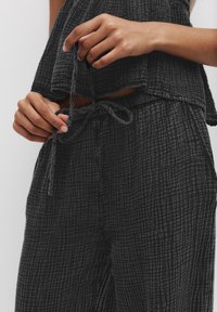 Set iz črnega teksturiranega blaga, ki vključuje ohlapen crop top in hlače na vezalko. Opozablja na šahovnico z rahlo gubanjem v teksturi.