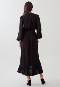 Robe maxi noire fluide avec de longues manches bouffantes, taille ceinturée et ourlet volants. Texture lisse et design minimaliste.
