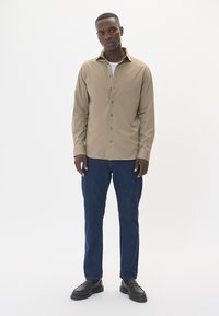 Chaki ilga rankovė marškiniai su mygtukų uždarymu, dėvimi ant baltos marškinėlės, derinami su tamsiai mėlynomis džinsinėmis kelnėmis ir juodais bateliais su raišteliais.