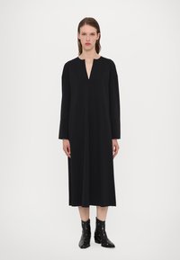 Black, long-sleeve dress with a V-neckline, made from a smooth fabric. It features a straight silhouette and falls to mid-calf. Black ankle boots.