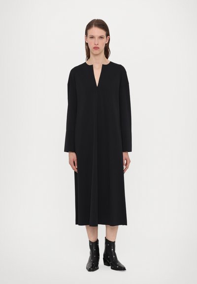 Crna haljina dugih rukava s V dekolteom, izrađena od glatke tkanine. Ima ravnu siluetu i doseže do sredine lista. Crne čizme do gležnjeva.