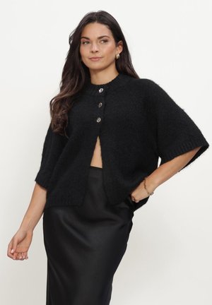 Vrouw met lang donker haar draagt een zwarte pluizige cardigan met drie knopen over een zwarte satijnen rok, staand tegen een effen lichte achtergrond.