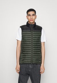 Chaleco acolchado con parte superior negra y parte inferior verde oscuro, que cuenta con costura horizontal, cuello alto y cierre de cremallera. Dos bolsillos laterales.