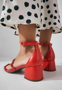 Røde læder sandaler med blokhæle, justerbare ankleremmer og åben tå design. Præsenteret på en baggrund af hvidt stof med prikker.