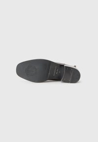 Semelle de chaussure en cuir noir avec une texture lisse, présentant un léger talon, un logo en relief et des clous en métal pour un détail supplémentaire.