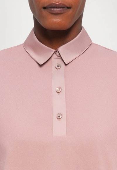 Lys rosa polo skjorte med en myk tekstur, krage, og fire knapper foran. Stoffet har et subtilt, vevet mønster.