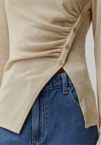 Beige Langarm-Oberteil mit gerüschter Seite und Knopfdetails, kombiniert mit blauen Jeans im Standard-Schnitt und passender Naht.