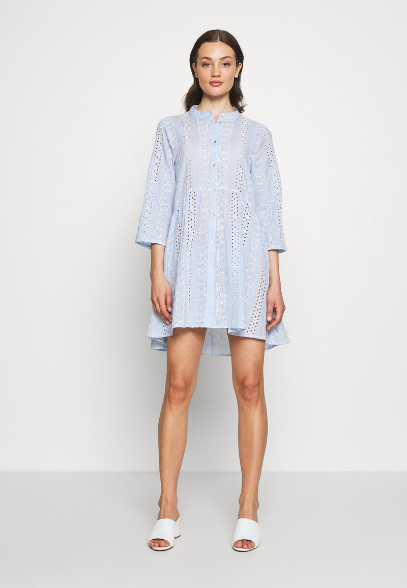 Vestido azul claro feito de algodão com detalhes em ilhós, apresentando uma frente com botões, colarinho e mangas de 3/4 de comprimento; silhueta semi-ajustada.