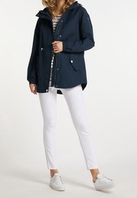 Mujer con una chaqueta con capucha azul marino, camiseta a rayas, pantalones ajustados blancos y zapatillas blancas, de pie contra un fondo liso.