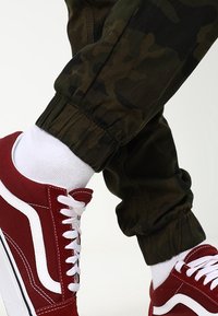 Červené plátěné tenisky s bílými tkaničkami a bočními pruhy, doplněné černými maskáčovými tepláky se elastickými manžetami a hladkou texturou.