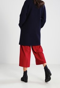 Cappotto lungo blu, pantaloni rossi a gamba larga e stivali neri alla caviglia. Il cappotto ha una vestibilità comoda; i pantaloni sono plissettati e di lunghezza corta.