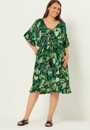 Kvinde smilende iført knælange grønne kjole med bladmønster, flæser på ærmer og underkant, parret med sorte sandaler, stående på lys baggrund.