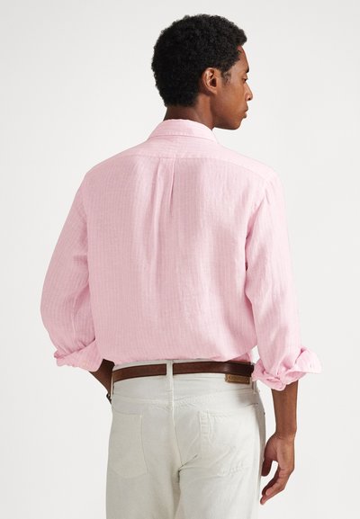 Muž otočený chrbtom, oblečený v svetloružovej košeli s dlhými rukávmi vyhrnutými až po lakte, bielych nohaviciach a hnedom opasku, na jednoduchom pozadí.