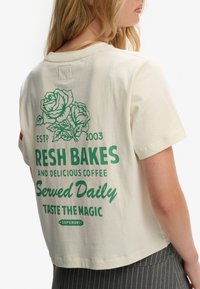 Cremefarvet bomulds-T-shirt med grøn trykt tekst og rosetryk på ryggen. Korte ærmer og afslappet pasform. Brandlogo synligt.