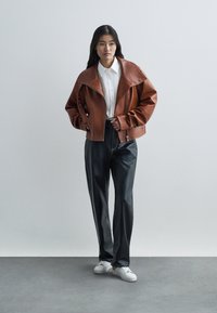 Brun lær cropped jakke med bred krage, hvit skjorte med knapper, svarte vide bukser og hvite sneakers mot en nøytral bakgrunn.