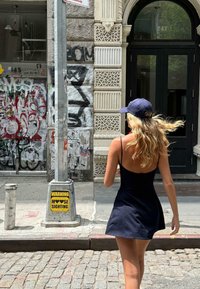 Robe noire à bretelles avec un ourlet évasé, portée avec une casquette de baseball bleu marine. Le cadre urbain présente des graffitis sur un bâtiment en briques et un panneau d'avertissement.