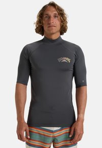 Grå surftröja med kort ärm och hög krage, med broderad logotyp på bröstet, tillverkad av stretchigt, fukttransporterande material.