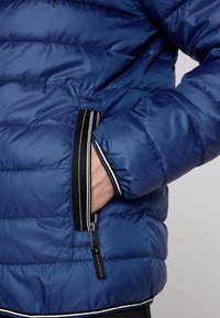 Giacca puffa blu con cuciture orizzontali, tasca laterale con zip e accenti neri. Materiale liscio e lucido con una texture morbida.