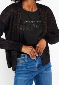 Svart, glittrig cardigan med öppen front, bärs över en svart skjorta med texten "just be HAPPY" i metallisk stil. Matchad med blå jeans.