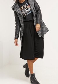 Schwarzer knielanger Rock kombiniert mit einem schwarzen T-Shirt mit grafischem Aufdruck und einem langen, strukturierten grauen Mantel. Schwarze Stiefeletten runden den Look ab.