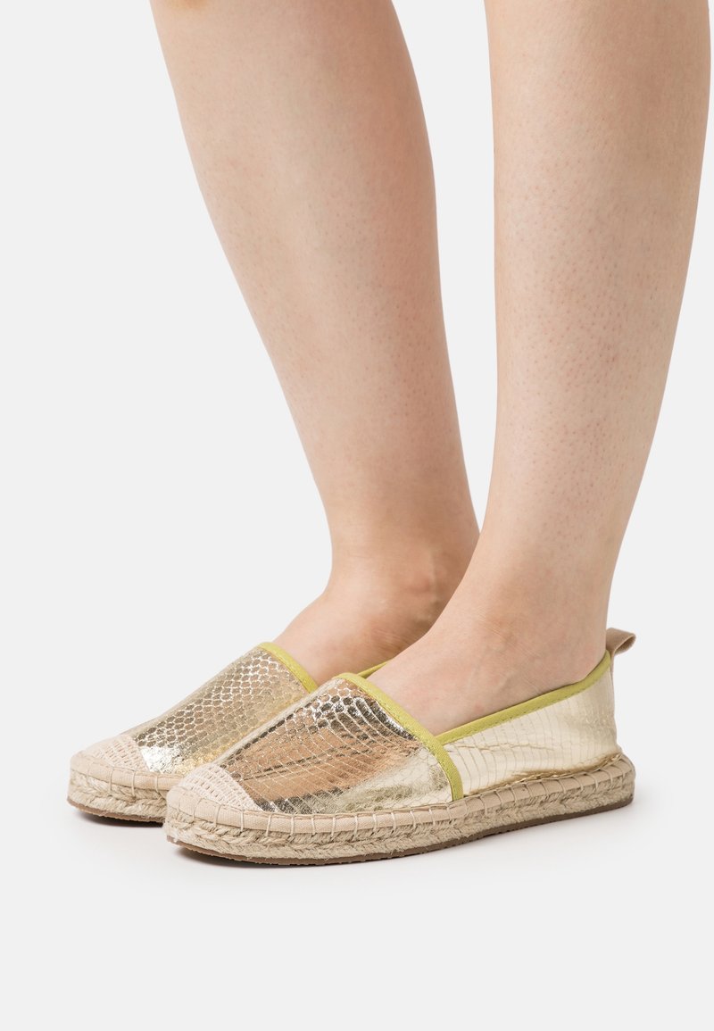 Pieds chaussés d’espadrilles slip-on métalliques dorées avec semelles en jute tressée sur fond blanc.