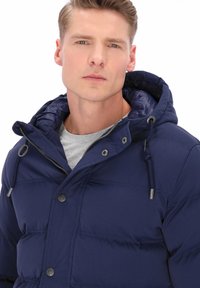 Mørkeblå dynejakke med hætte, lynlås og trykknapper. Har vandrette syninger og snoretrækdetaljer omkring hætten.