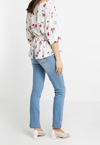 Blusa floreale con maniche a sbuffo, vita cintrata, sfondo bianco e motivi di fiori rossi. Abbinata a jeans azzurri chiari e scarpe con tacco bianche.