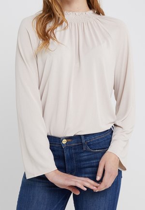 Mujer vistiendo una blusa de manga larga, beige claro, con escote fruncido metida dentro de unos vaqueros azules, manos entrelazadas suavemente al frente.