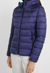 Giacca imbottita blu navy con cappuccio, caratterizzata da una texture trapuntata, chiusura frontale con cerniera e due tasche laterali con cerniera. Fodera interna a contrasto.