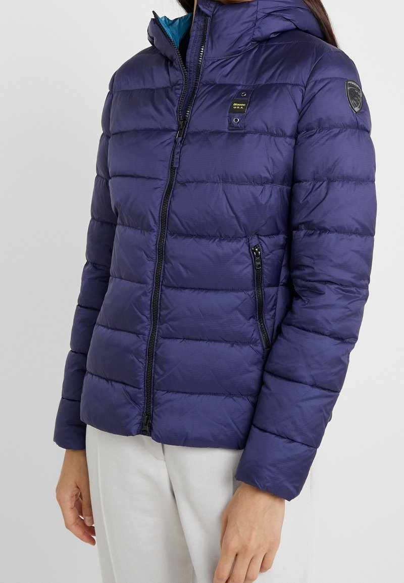 Giacca imbottita blu navy con cappuccio, caratterizzata da una texture trapuntata, chiusura frontale con cerniera e due tasche laterali con cerniera. Fodera interna a contrasto.