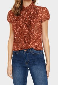 Rostfärgad, semi-transparent blus med volangdetaljer och vita prickar, korta ärmar och hög krage, parat med blå jeans.