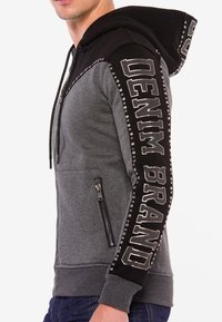Grauer und schwarzer Kapuzenpullover aus weichem Material; verfügt über eine zipptasche, auffällige Aufschrift auf den Ärmeln und Nietenakzente entlang der Nähte.