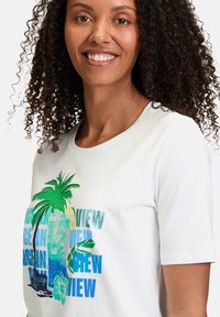 T-shirt blanc à manches courtes avec un imprimé tropical coloré représentant un palmier, un verre et des éléments textuels en bleu et vert.