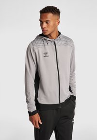 Sudadera con cremallera gris que presenta una sección superior texturizada con puntos, detalles en negro en los laterales y una capucha con cordón. Incluye un logo en el pecho.