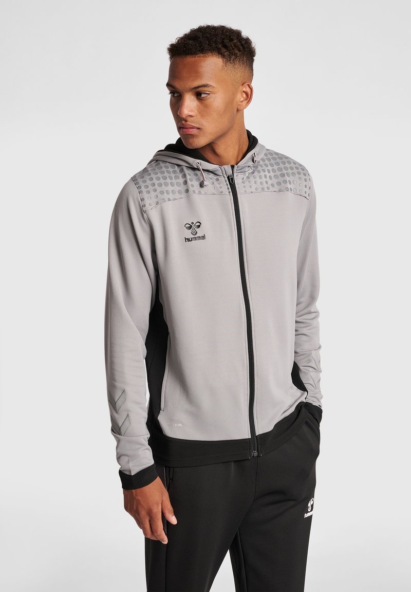 Sudadera con cremallera gris que presenta una sección superior texturizada con puntos, detalles en negro en los laterales y una capucha con cordón. Incluye un logo en el pecho.