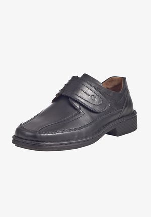 Black leather shoe with a rounded toe, stitched detailing, and a Velcro strap across the instep. Low, textured rubber sole.