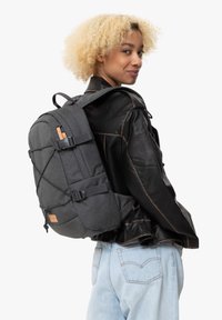 Sac à dos gris en tissu, doté de bretelles ajustables, de sangles frontales et d'un accent en cuir. Conçu avec une forme structurée et des poches latérales.