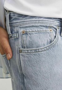 Lichtblauwe denim jeans met een close-up van de zak met zichtbare stiksels en klinknagels. De stof heeft een lichte textuur en patroon.