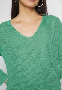 Femme portant un pull en V en tricot vert et de grandes boucles d'oreilles en or, avec de longs cheveux noirs et une main sur la hanche.