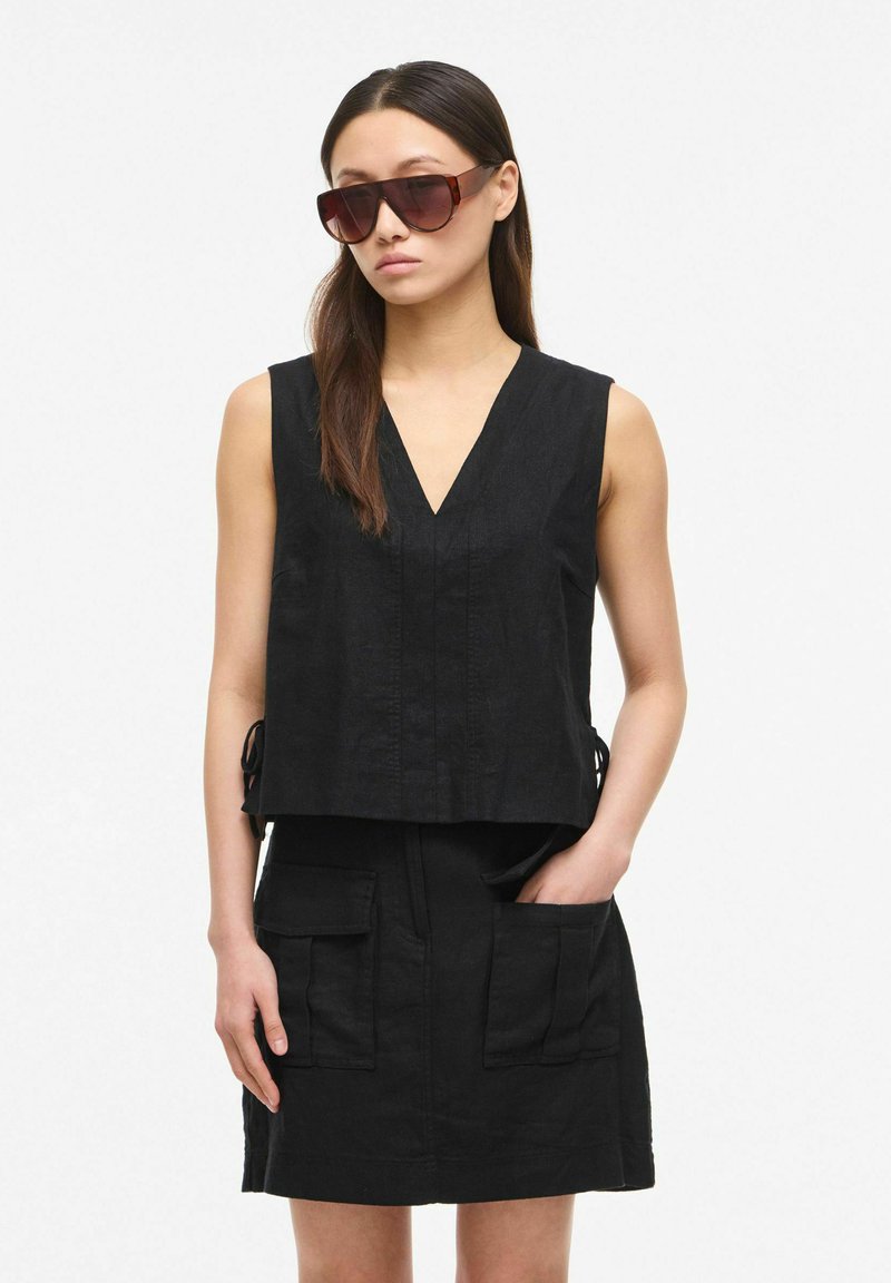 Femme portant de grandes lunettes de soleil marron, un haut noir sans manches à col en V et une jupe noire avec de grandes poches devant, debout avec une main dans la poche.