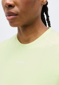 Κοντινή λήψη του κάτω μέρους του προσώπου, του λαιμού και του άνω μέρους του θώρακα ενός ατόμου που φοράει ανοιχτοπράσινο πουκάμισο με λαιμόκοψη τύπου crewneck με μικρό λευκό λογότυπο.