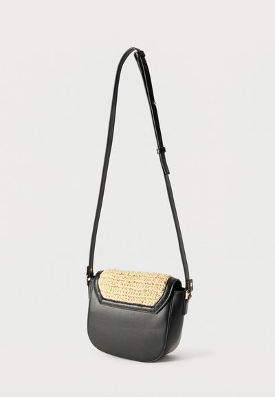 Μαύρο δερμάτινο τσαντάκι crossbody με ρυθμιζόμενο λουράκι και πλεκτό μπεζ καπάκι, απεικονιζόμενο με φόντο απλή ανοιχτή επιφάνεια.