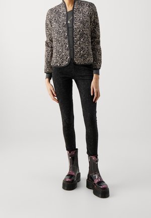 Personne portant une veste à motifs, un pantalon foncé ajusté et des bottes à plateforme violettes lacées, debout devant un fond uni.
