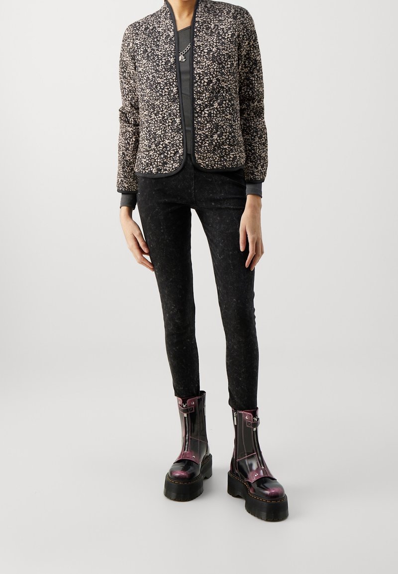 Personne portant une veste à motifs, un pantalon foncé ajusté et des bottes à plateforme violettes lacées, debout devant un fond uni.