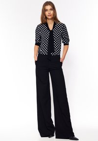 Czarny i biały sweter w kropki z długimi rękawami i zawiązanym przodem, w połączeniu z szerokimi czarnymi spodniami o gładkiej fakturze i dopasowanym kroju.