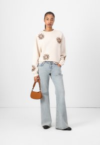 Cream sweater with sun-pattern embroidery, paired with light blue flared jeans. A small brown handbag completes the outfit. Black footwear visible.
