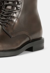 Stivaletto in pelle marrone con punta tonda, cuciture rinforzate e lacci neri. Presenta un tacco a blocco nero, basso e robusto. Texture liscia e lucida.