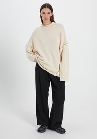 Suéter de punto beige con un cuello alto, ajuste holgado y detalles de corte. Combinado con pantalones de pierna ancha negros y zapatillas oscuras.