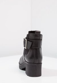 Botín de cuero negro hasta el tobillo con tacón cuadrado, panel elástico lateral, detalle de hebilla prominente y acabado en cuero texturizado.