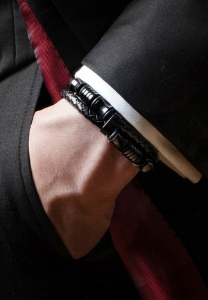 Black beaded bracelet with various textures and shapes, worn on a wrist, paired with a suit jacket and a contrasting red tie.